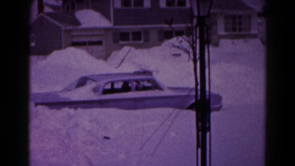 A grainy 8mm frame shows the snowbound sedan lower in the frame, with residential buildings and vertical supports nearby.