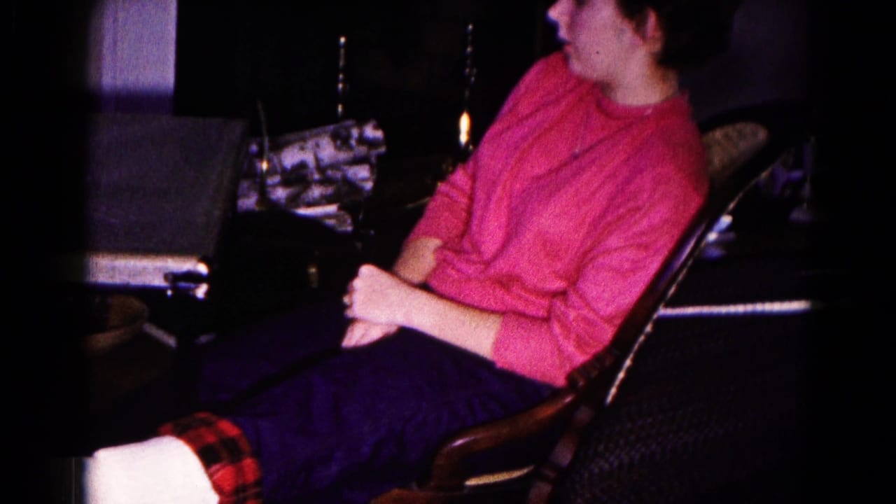 In a 1959 Hagerstown home-movie frame, a woman in a pink sweater sits near a card table in a dim living room.