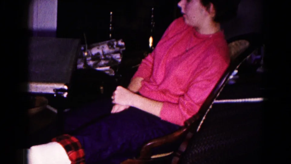 In a 1959 Hagerstown home-movie frame, a woman in a pink sweater sits near a card table in a dim living room.