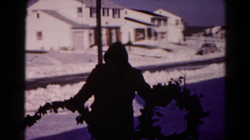A hooded adult in 1959 Hagerstown holds evergreen garland beside a wreath with snowbanks and houses in the background.