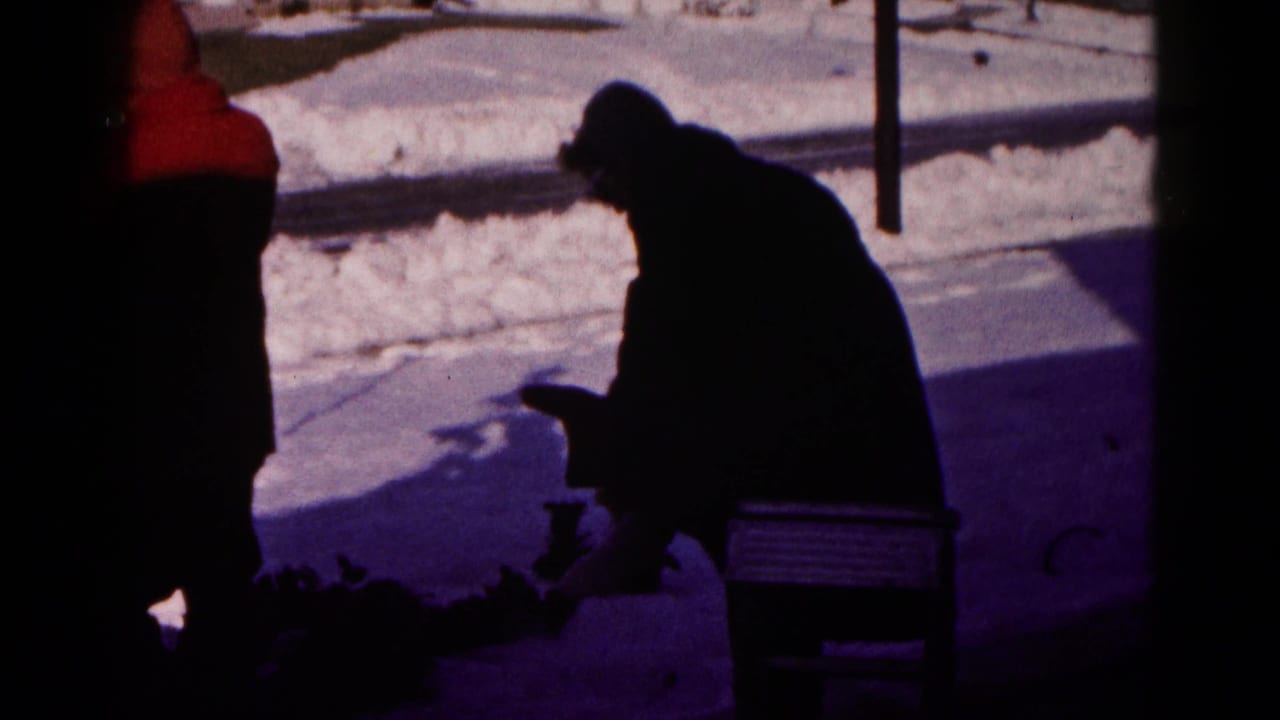 In a 1959 Hagerstown home-movie frame, a man sits on a bench in a snowy yard while a bundled figure stands at the left edge.