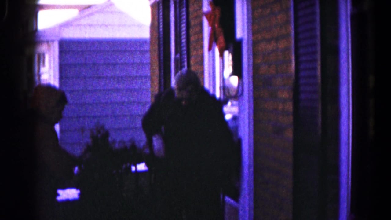 Two people sit in silhouette on a Hagerstown porch in 1959 8mm footage, with brick siding and a neighboring house visible.