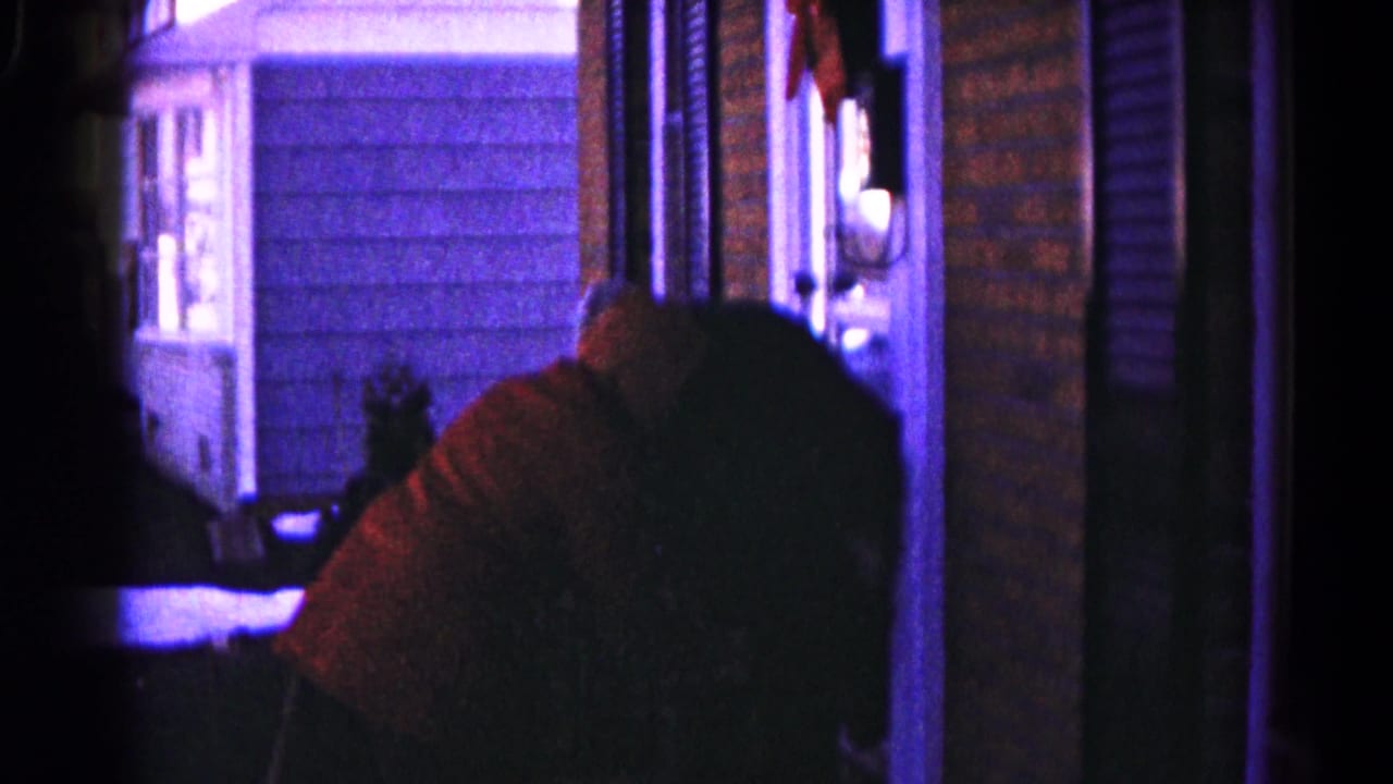 A hooded figure leans near another person on a dim porch beside brick siding.