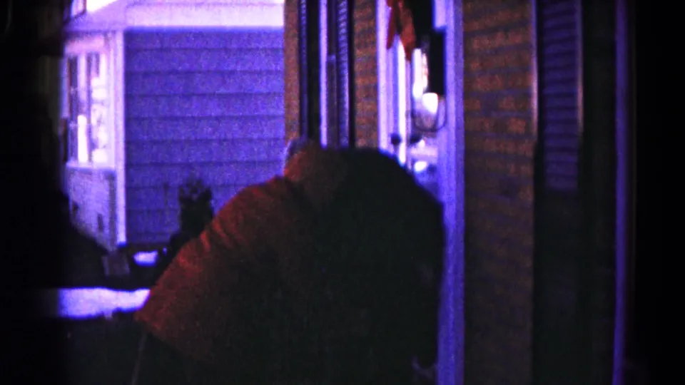 A hooded person and another dark-clothed figure sit close together on a dim Hagerstown porch in 1959 home-movie footage.
