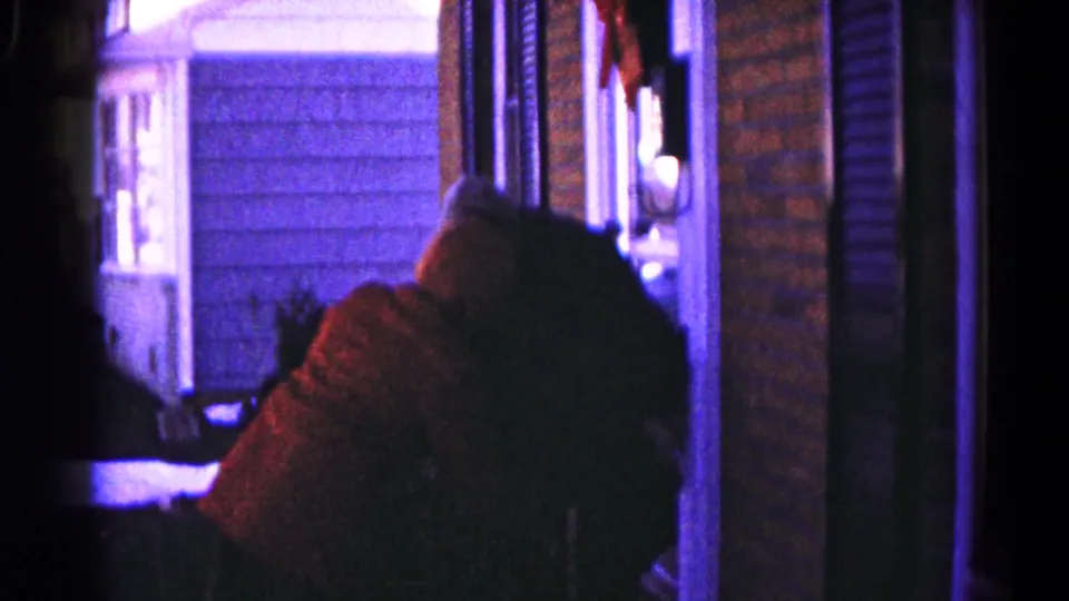 A person in a reddish garment leans forward beside another figure on a low-lit porch in 1959 Hagerstown home-movie footage.