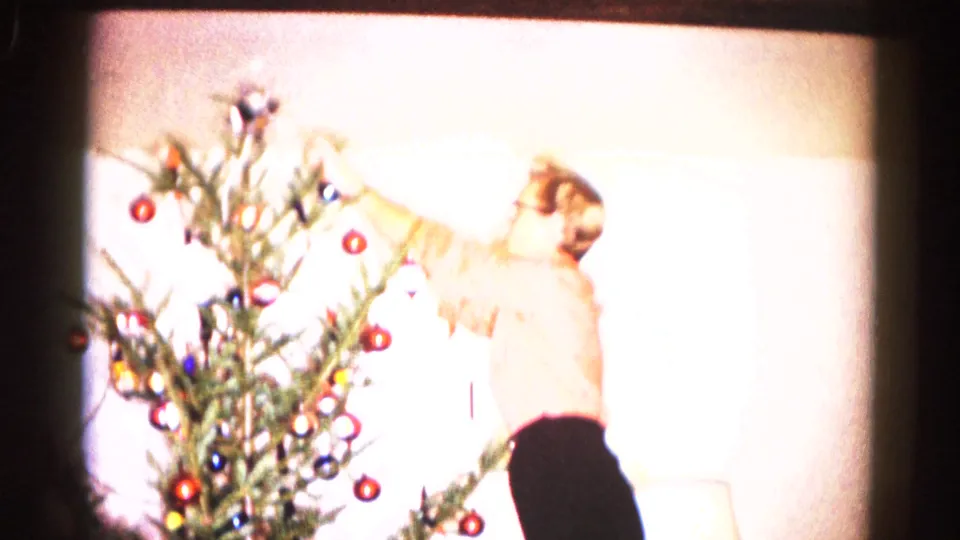 In this 1959 Hagerstown home-movie frame, a person reaches toward a small Christmas tree with round ornaments in a bright living room.