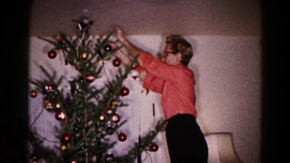 A 1959 Hagerstown interior frame shows a person in a red sweater adding ornaments to a small Christmas tree beside a lamp.