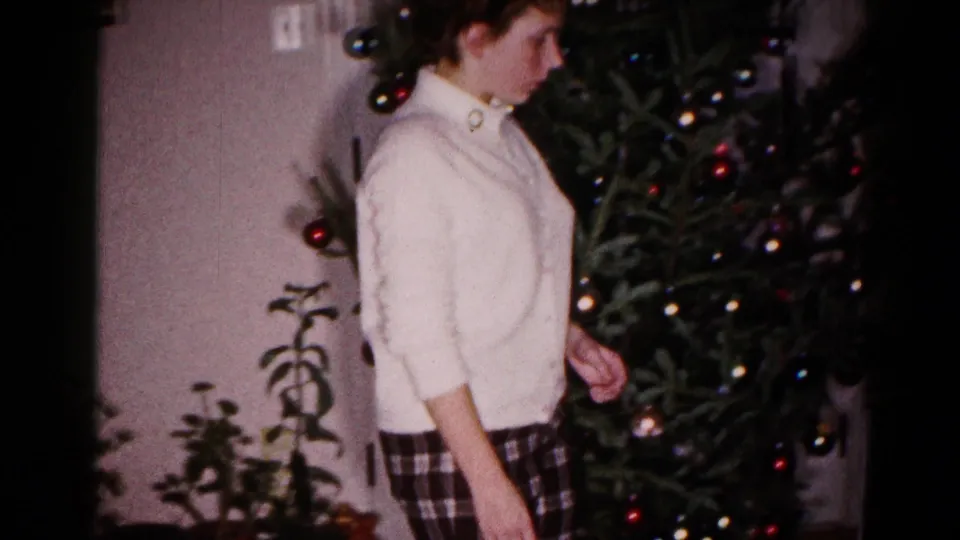 A close home-movie view shows the young woman near the decorated Christmas tree, with ornaments and interior plants in the frame.
