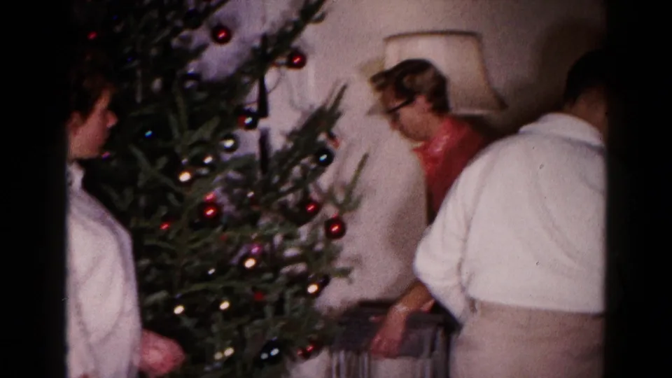 Two adult men and the young woman gather at the Christmas tree in the Hagerstown home, with a lamp and ornaments visible.
