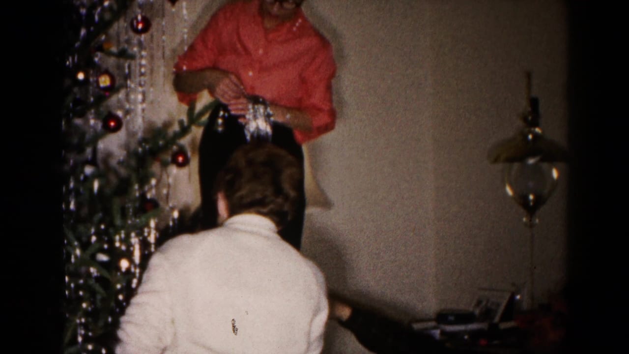 Two women decorate a Christmas tree indoors with silver tinsel and ornaments.