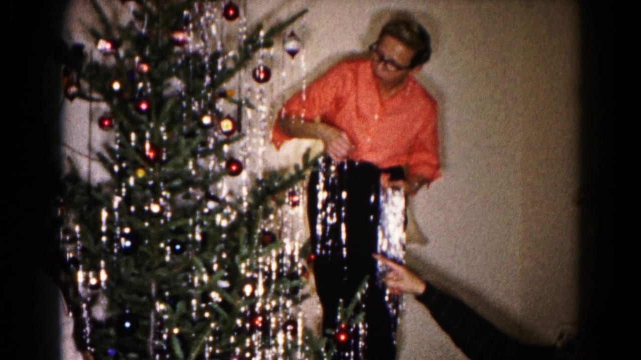 The man adjusts hanging tinsel on the Christmas tree while a second person's hand points toward an ornament or branch.