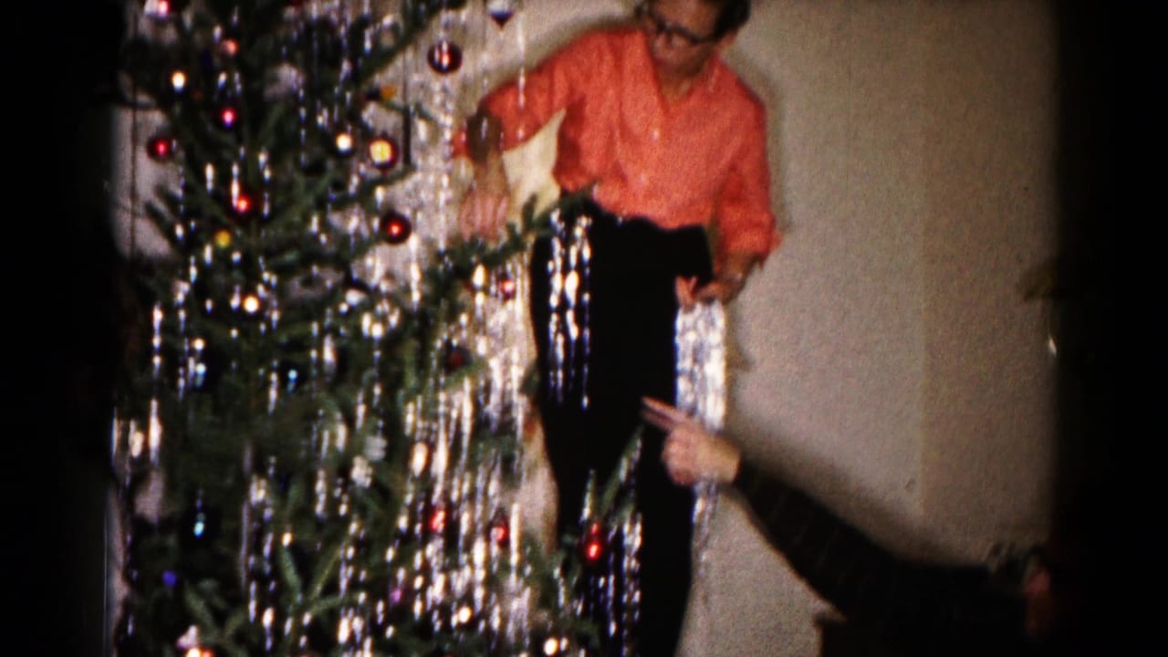 In the home interior, the man stands near the decorated tree with long silver tinsel strands hanging as a hand points from the lower right.