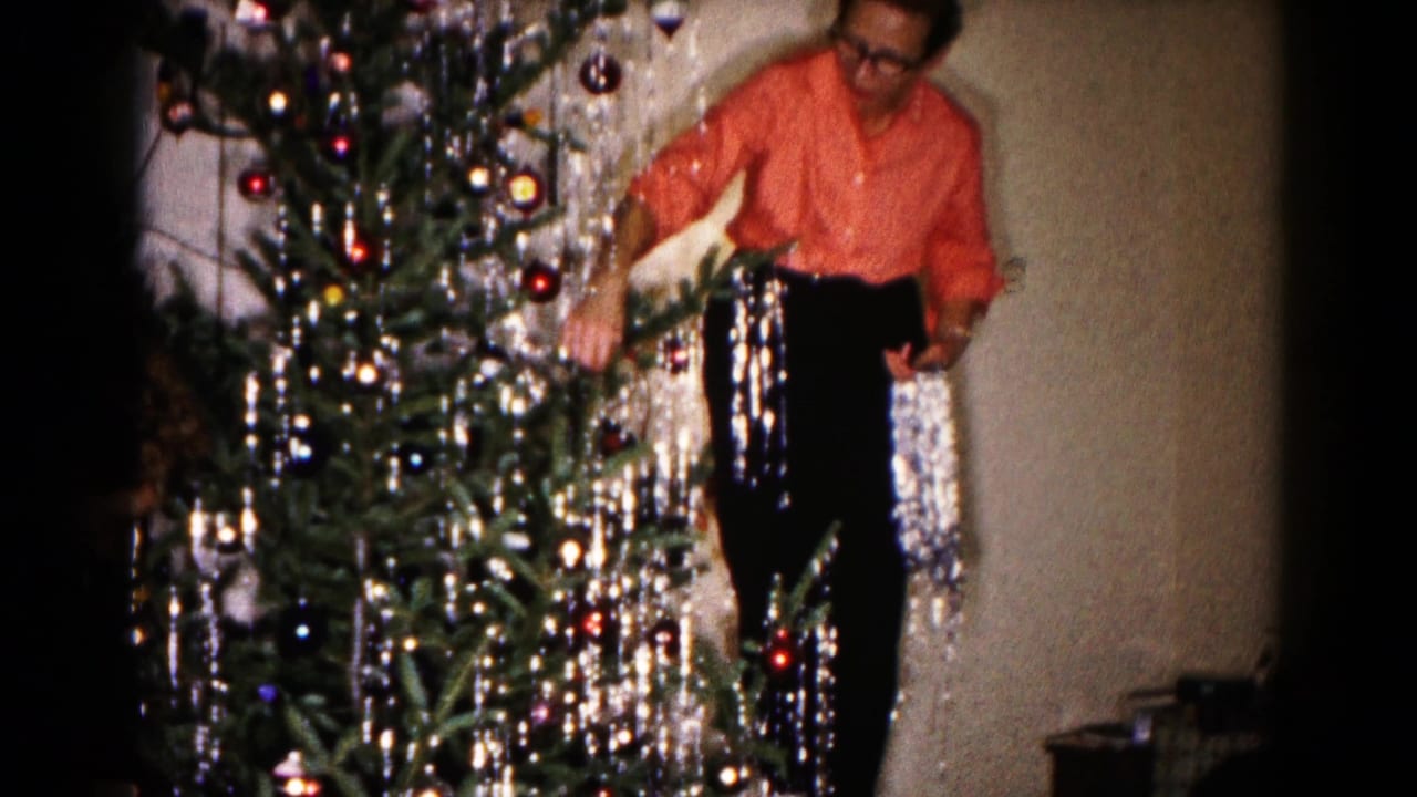 In a 1959 Hagerstown home-movie frame, a woman in a coral blouse stands beside a Christmas tree with silver tinsel and ornaments.