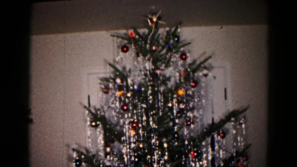 The 1959 home-movie frame centers on a Christmas tree with hanging silver tinsel, colored ornaments and a star near the top.