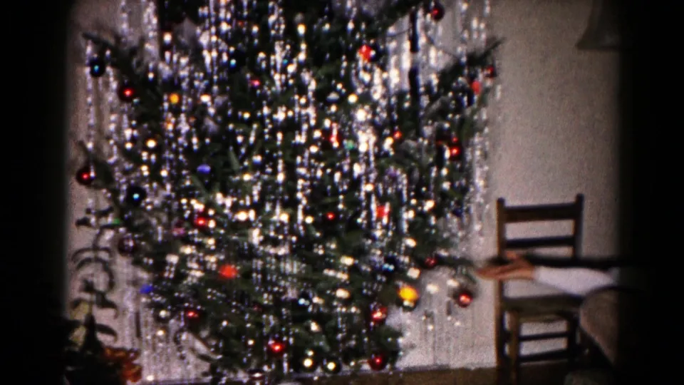 1959 Hagerstown home-movie frame of a tinsel-covered Christmas tree in a private room, with a chair and partial upholstered furniture visible.