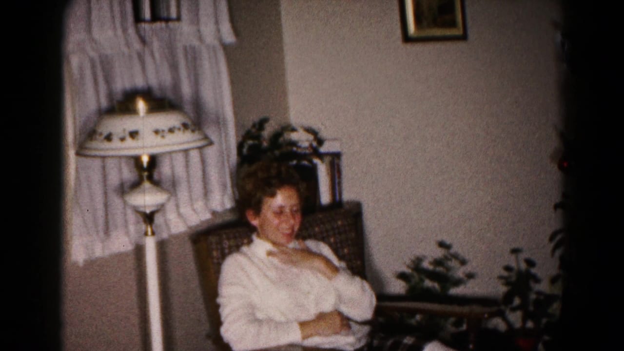 A woman in a white sweater sits smiling in a living-room chair, with a floor lamp, curtains, and plants behind her.