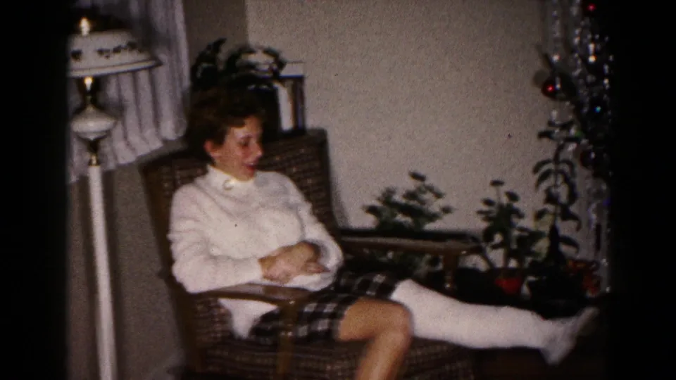 The seated woman looks down as her casted leg extends from the armchair in a softly lit 1959 home interior.