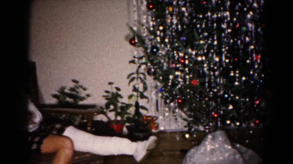 A casted leg extends from an armchair beside a decorated Christmas tree with ornaments, tinsel-like strands, plants, and a wall behind it.