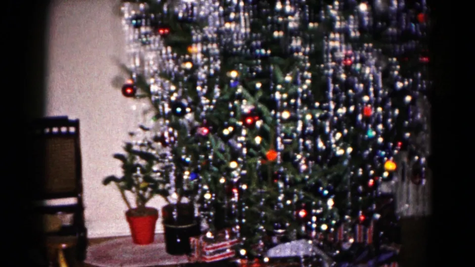 1959 Hagerstown home-movie frame showing a Christmas tree with silver tinsel, ornaments, potted plants, presents, and a wooden chair.
