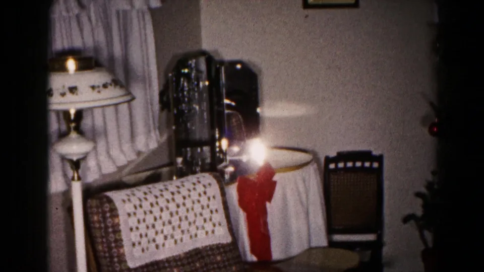 1959 Hagerstown living room frame with lamp glare, curtains, patterned sofa, crocheted throw, white table covering with red bow, chair, and plant.