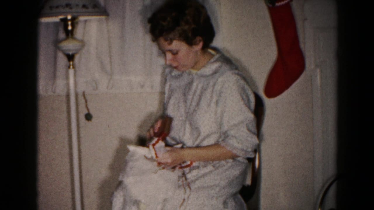 Woman in a light nightgown sits by a floor lamp while opening a wrapped Christmas present.