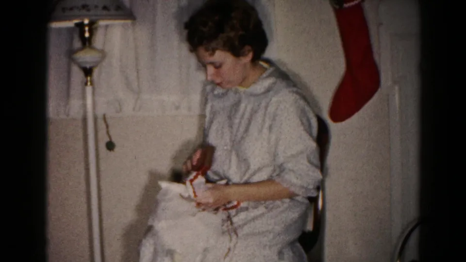 In a 1959 Hagerstown home-movie frame, a seated woman in a light nightgown unwraps a present beside a floor lamp and red stocking.