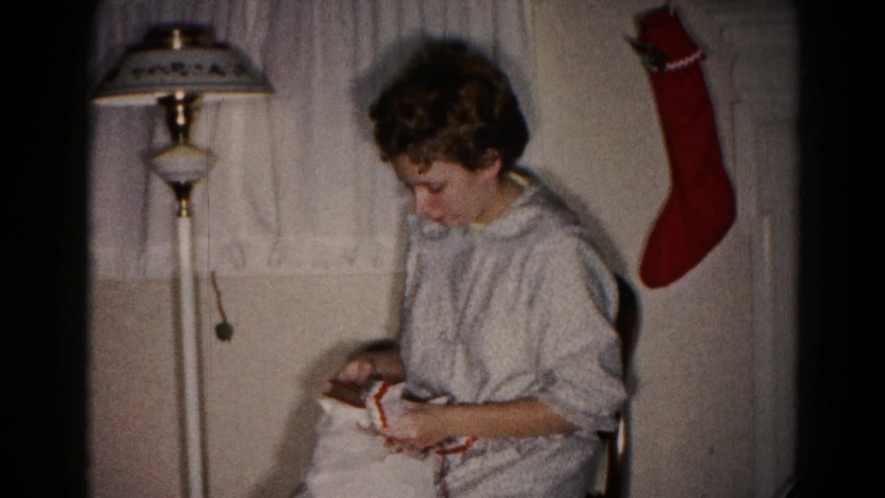 The woman continues unwrapping the Christmas gift in a private Hagerstown interior, with a patterned lamp shade and red stocking behind her.
