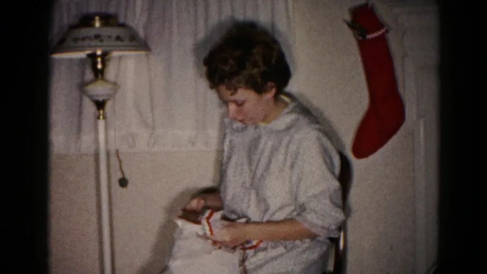 The woman continues unwrapping the Christmas gift in a private Hagerstown interior, with a patterned lamp shade and red stocking behind her.