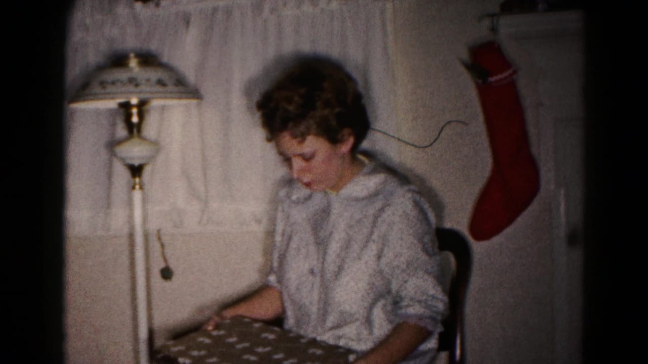In a 1959 Hagerstown home interior, a seated woman looks down at a wrapped Christmas gift near a floor lamp and red stocking.