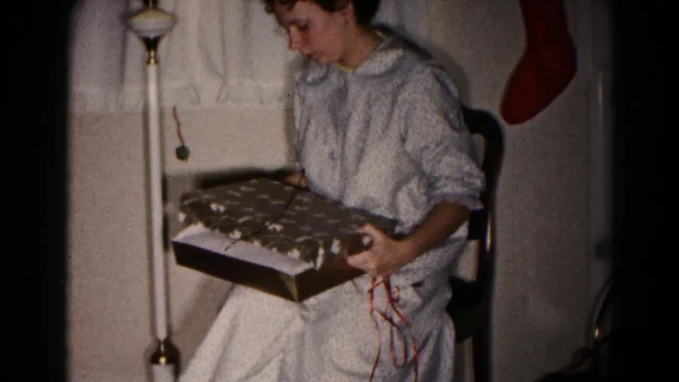 A seated woman in a light nightgown holds an opened Christmas package in the Hagerstown home-movie interior.
