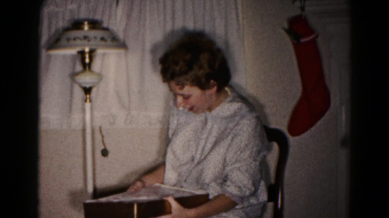 Young woman seated beside a floor lamp, looking down at a gift box in a home interior.