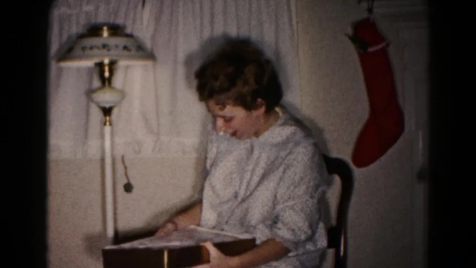 In 1959 Hagerstown home-movie footage, a young woman sits beside a floor lamp and looks into a gift box near a red stocking.