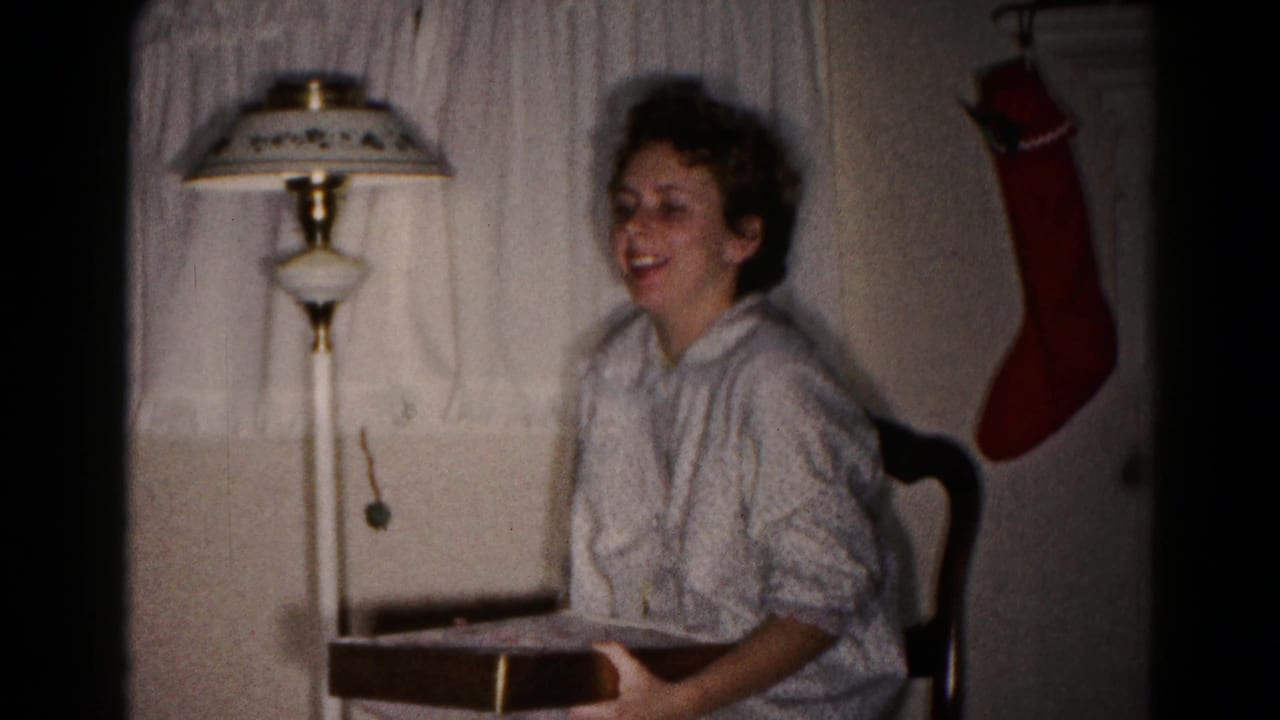 Young woman smiling while seated with an open gift box beside a lamp and wall stocking.