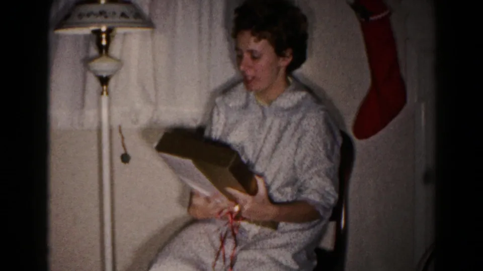 The woman holds the gift box and lid, with red ribbon visible on her lap in the 1959 Christmas home-movie scene.