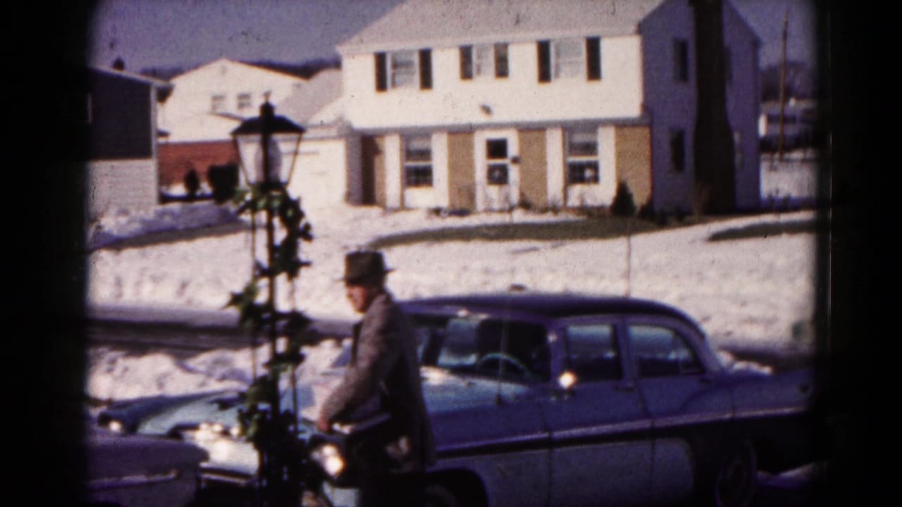 Older man in a fedora walks near a blue sedan on a snowy suburban street.
