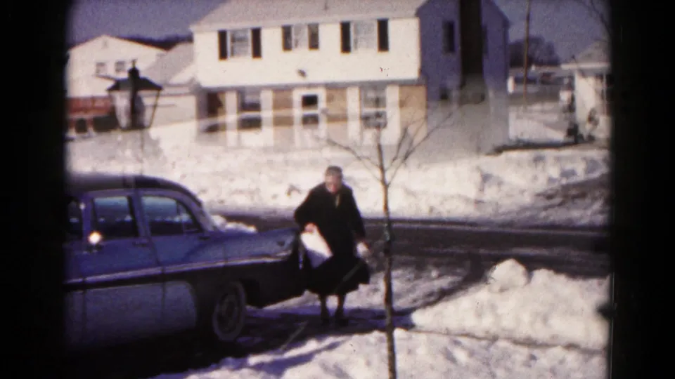 In the snowy Hagerstown residential scene, an older woman in a dark coat steps away from a blue sedan while carrying light-colored items.