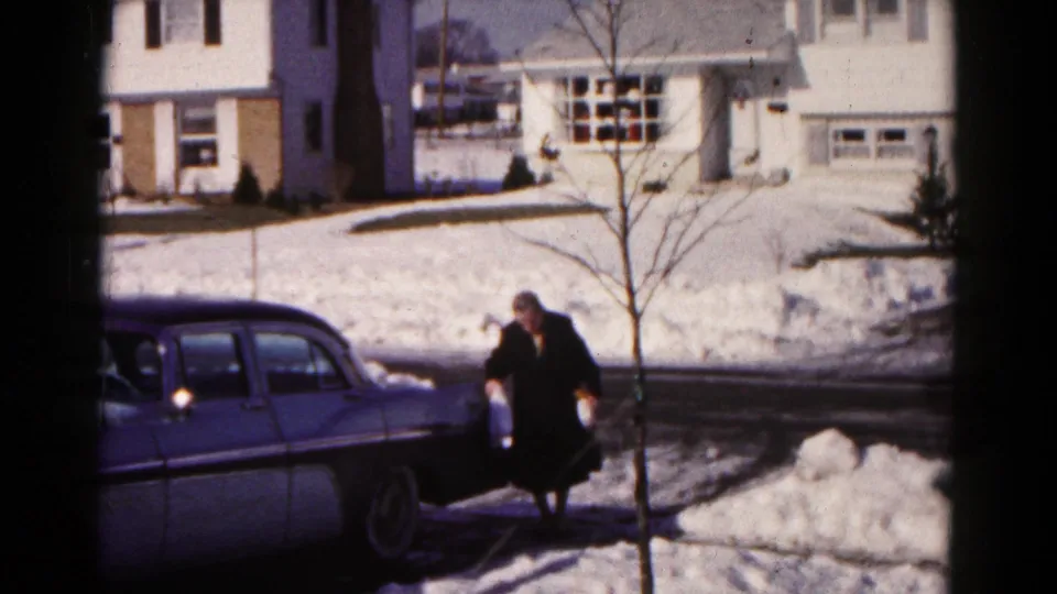 The older woman continues walking carefully beside the blue sedan in a snow-covered Hagerstown suburban neighborhood.
