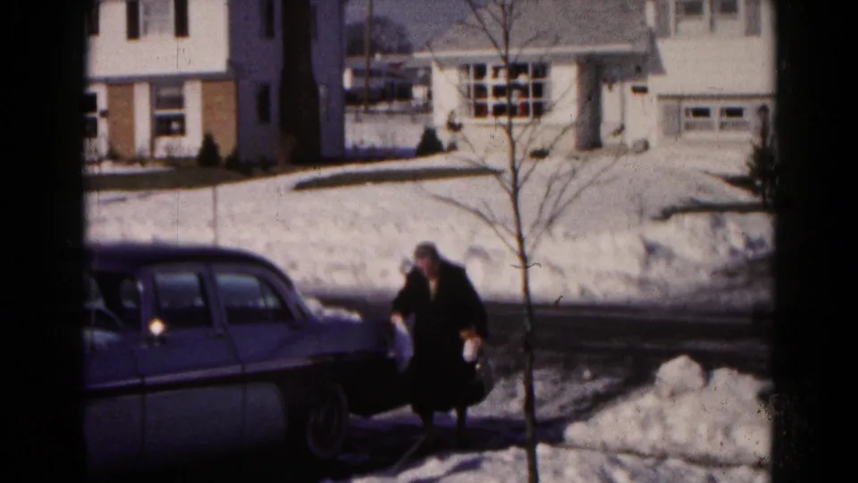 An older woman in a dark winter coat walks away from the parked blue sedan across the icy edge of a snowy Hagerstown driveway.
