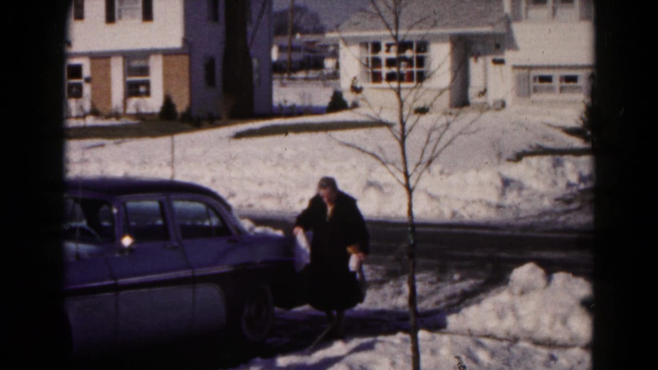 Woman in a dark coat walks through snow beside a parked two-tone sedan.