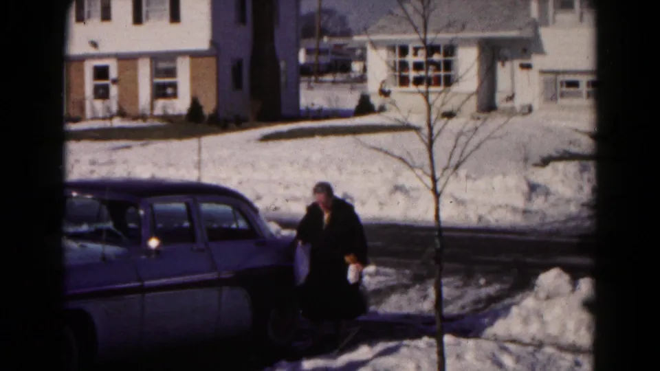 The woman continues across the snowy Hagerstown street area toward a two-tone sedan, with houses and bare trees behind her.