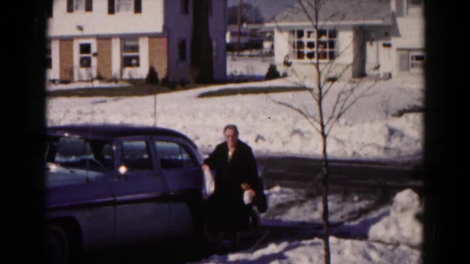 A 1959 Hagerstown home-movie frame shows the woman near the parked sedan, facing toward the camera amid deep snow and winter glare.