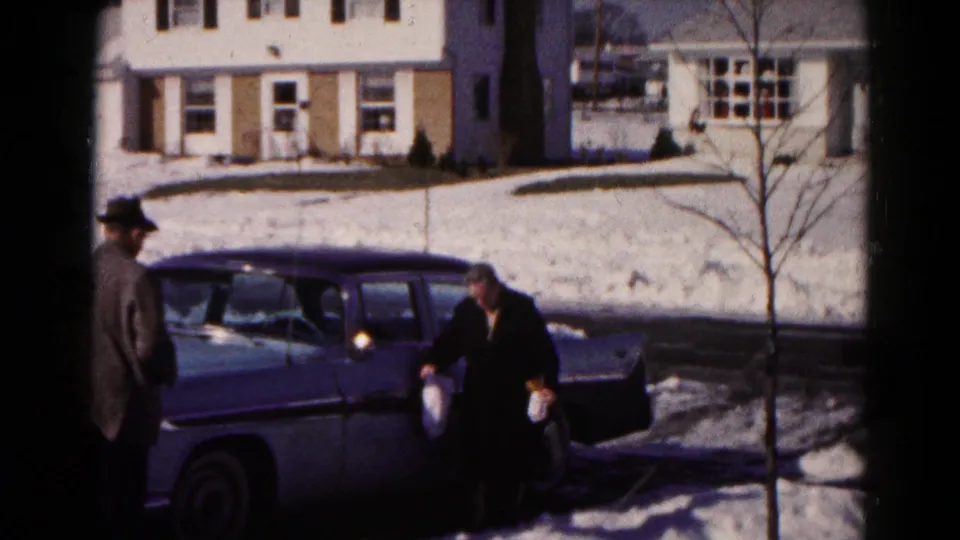 An adult man in a hat enters the frame while the woman stands beside the two-tone sedan on a snowy Hagerstown residential street.