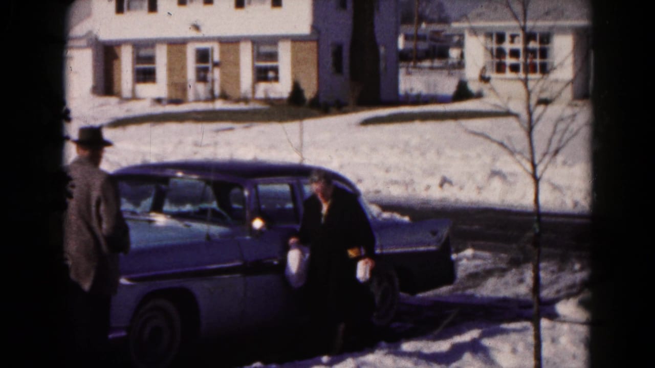 Two adults beside a classic sedan on a snowy residential street.
