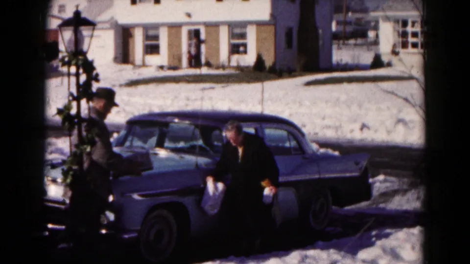 The woman appears to hold a grocery bag near the sedan while the man stands close by on the snowy residential street.