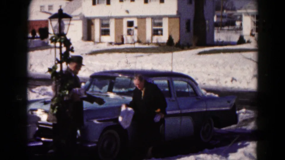 A woman with a grocery bag stands at the car while a man in a fedora remains nearby, framed by snow and suburban houses.