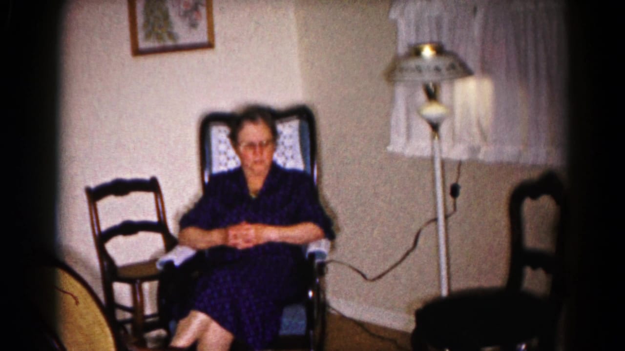 In a Hagerstown, Maryland home movie frame from 1959, a woman in glasses sits with folded hands in a living room chair.