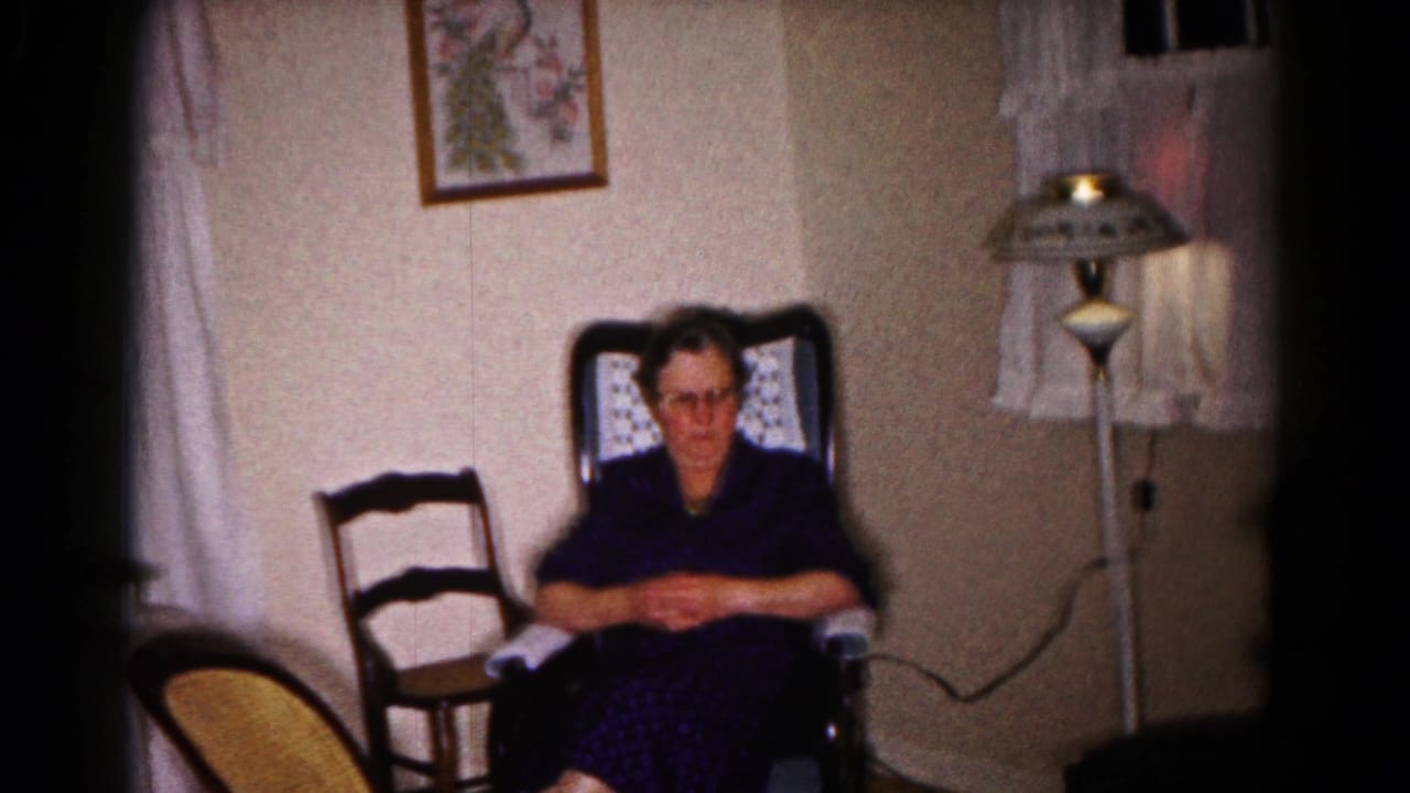 Seated woman faces the camera in a living room with chairs, curtains, and a lamp.