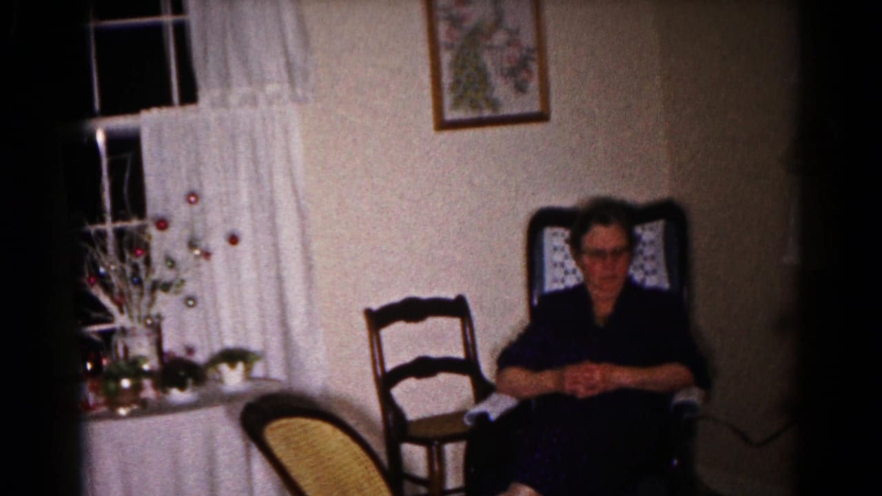 A wider 1959 Hagerstown home-movie frame shows the woman seated near curtained windows, a table display, and wooden chairs.