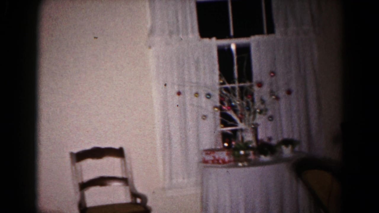 1959 Hagerstown home-movie frame showing a dim private room with white curtains, a window, a wooden chair, and a small holiday display on a table.