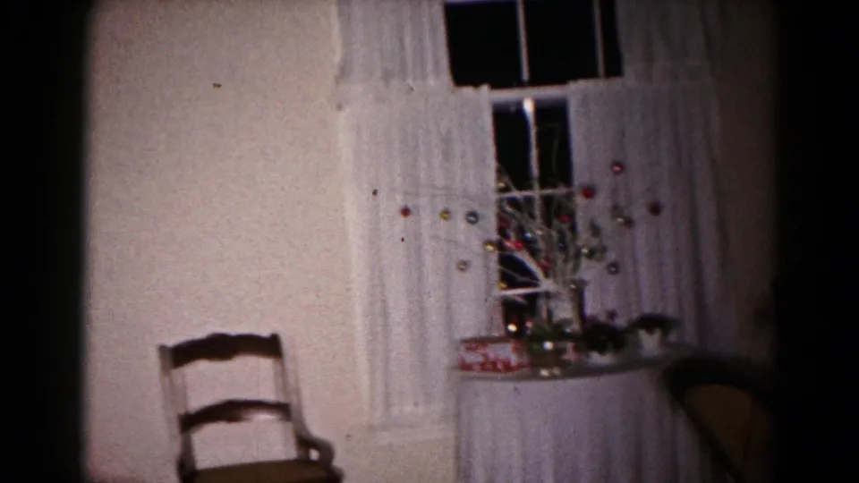 1959 Hagerstown home-movie frame showing a dim private room with white curtains, a window, a wooden chair, and a small holiday display on a table.